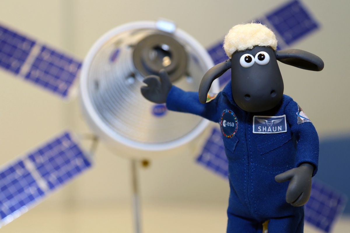 A plush cartoon sheep holds a hoof towards a small model of an Orion capsule.